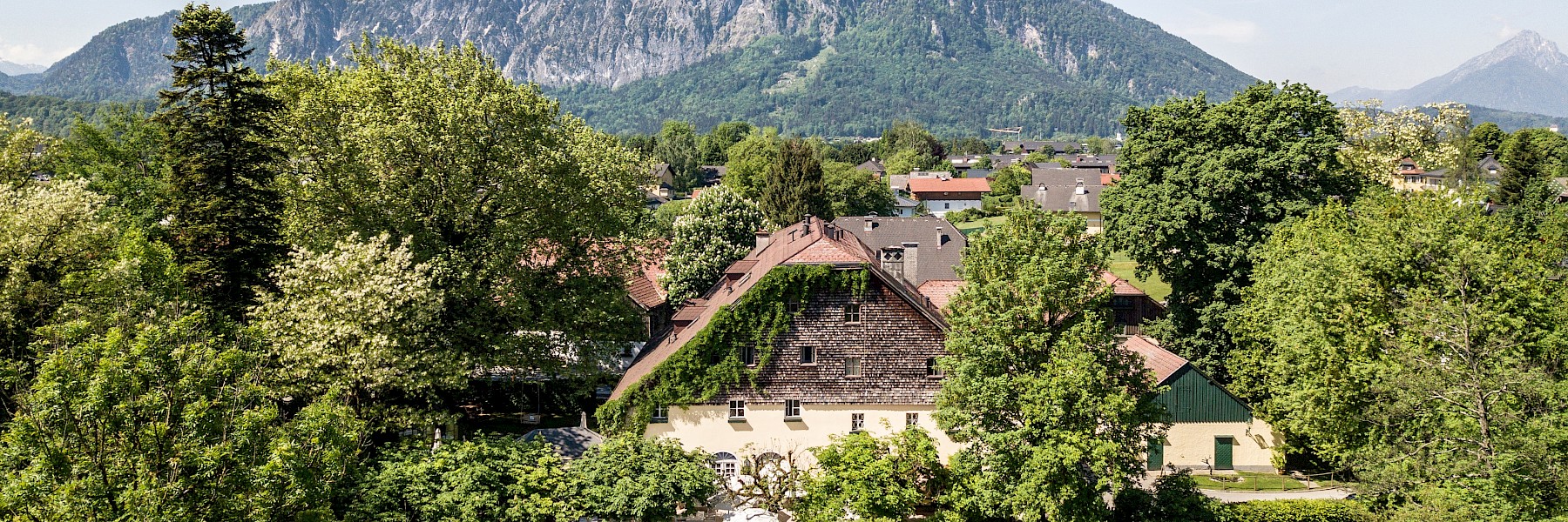 The image shows the Hotel Schlosswirt zu Anif surrounded by green trees. In the background, the Untersberg mountain rises majestically.