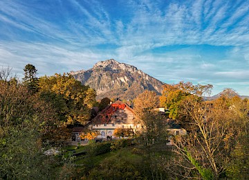 Landscape scene (c) Gassner Gastronomie, Foto Hechtfisch: The image shows an impressive drone view of Schlosswirt zu Anif surrounded by an autumn landscape. The historic building with its traditional wooden façade is nestled among colorful trees in warm autumn tones. In the background, the majestic Untersberg rises beneath a wide blue sky with delicate clouds. The garden area around the house appears peaceful, spacious, and beautifully maintained. Warm sunlight bathes the scenery in an inviting atmosphere. The image conveys a close connection to nature, tranquility, and exclusivity. Schlosswirt zu Anif appears as a special retreat for enjoyment, relaxation, and stylish stays near the city of Salzburg.