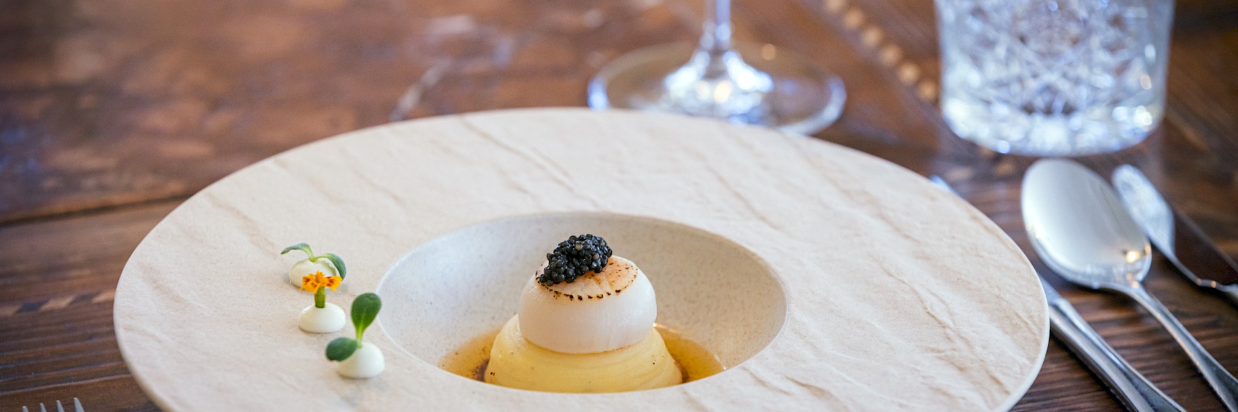 The image shows a beautifully plated gourmet dish served in a rustic, light-colored ceramic bowl placed on a wooden table. At the center is a perfectly seared fillet of fish with crispy skin, resting atop fresh handmade pasta. The dish is garnished with green asparagus, delicate zucchini ribbons, edible flowers, and microgreens. A light foam sauce surrounds the ingredients, adding a modern and elegant touch. The presentation is both refined and natural, reflecting freshness, high quality, and culinary artistry—perfect for a fine dining experience.